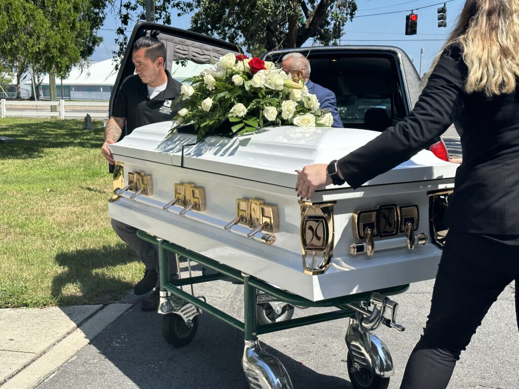 A white casket with white and red roses on top outdoors being rolled from a hearse. 