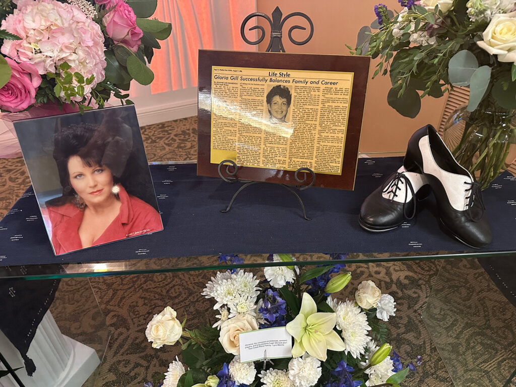 A picture, newspaper article, and black and white tap shoes resting on a table surrounded by floral arrangements. 