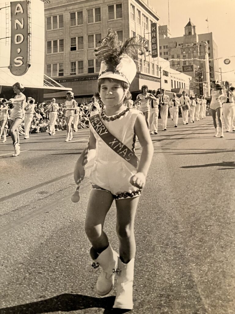 A black and white image of a child wearing a white hat with a large feather, a white costume with sequin trim, and white boots carrying a baton while walking in a parade. 