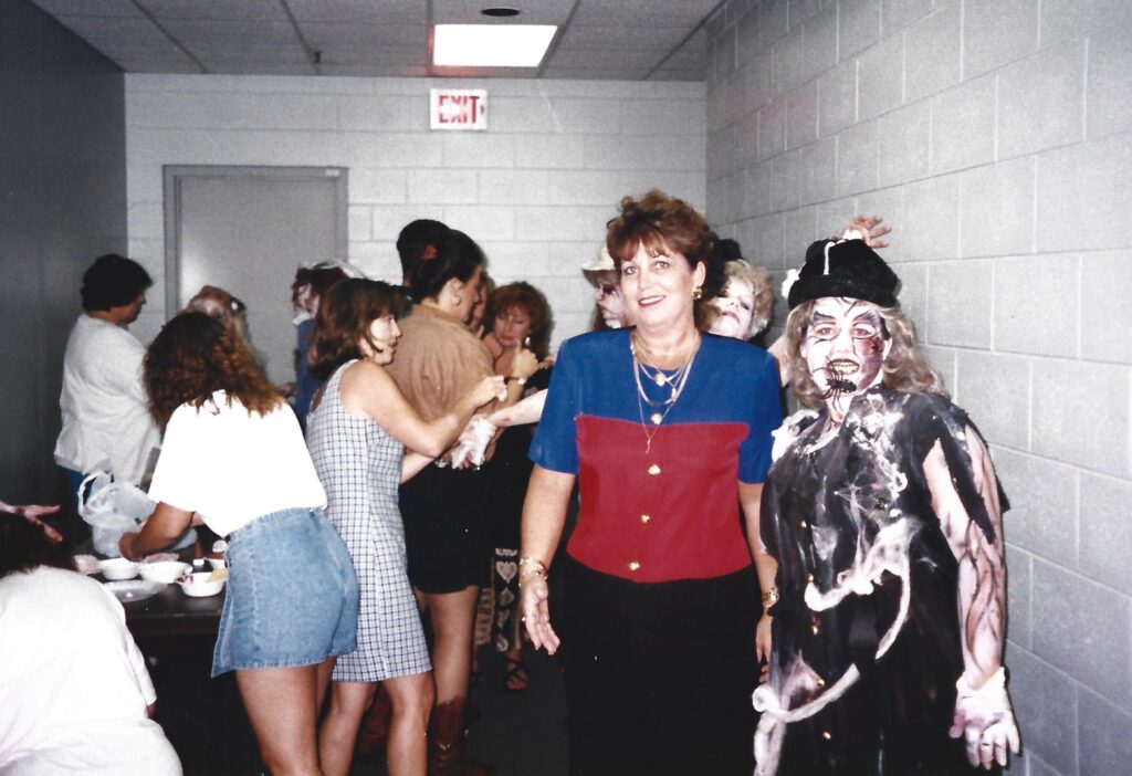 A woman smiling wearing a blue, red, and black shirt in front of a large group of people in front of a pale gray cement wall. 