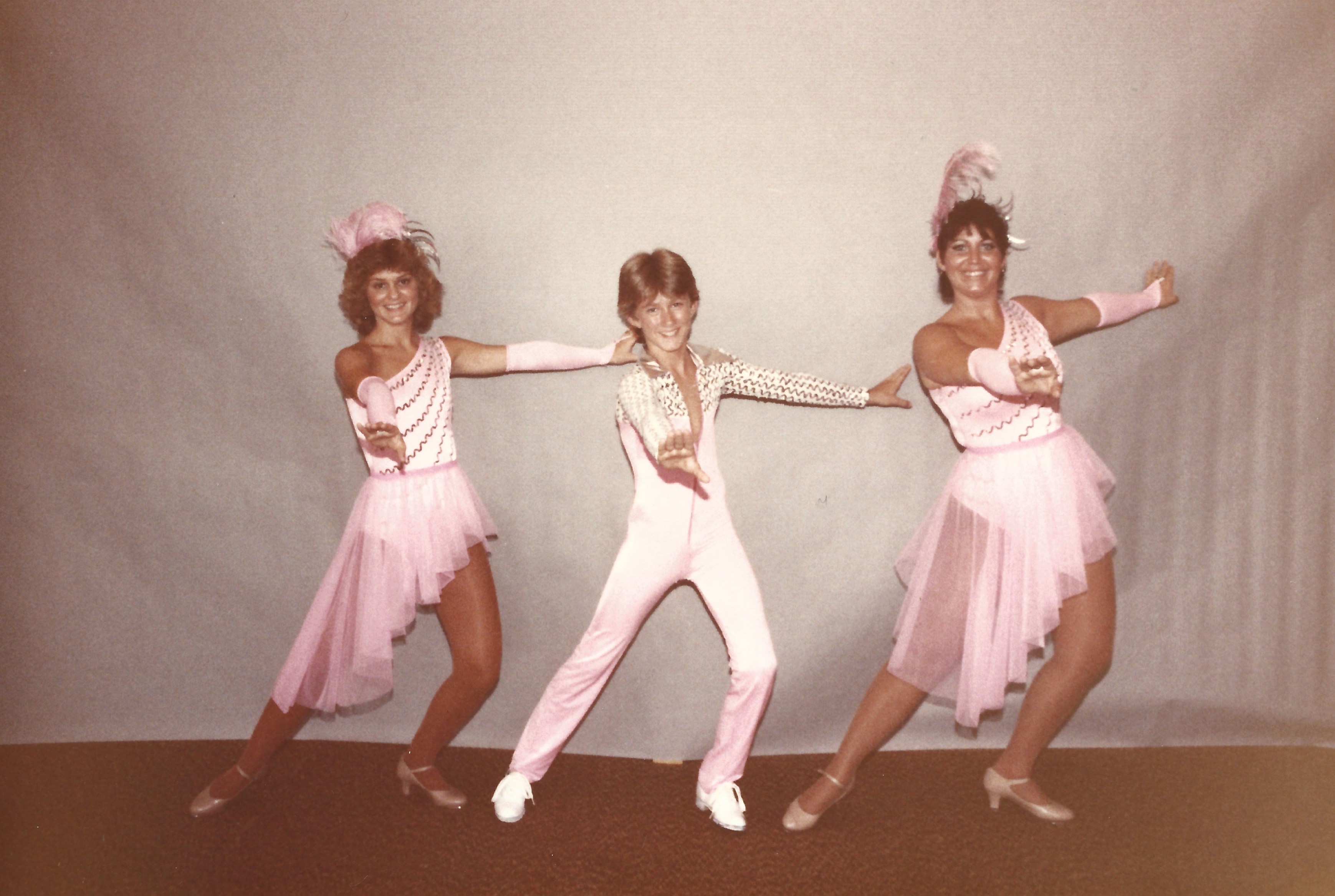 Three people posing in front of a beige background in pink and white costumes.