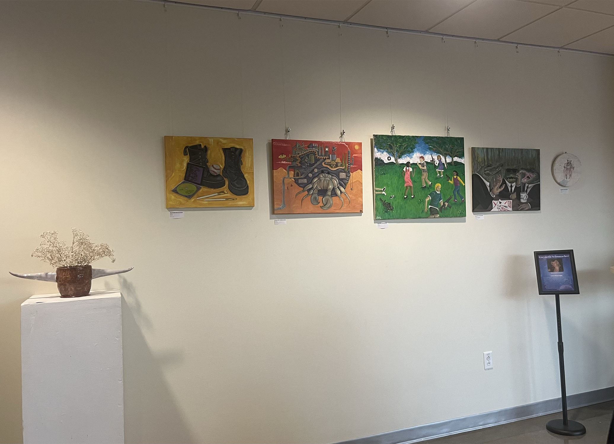 4 colorful oil paintings, 1 watercolor paining and 1 ceramic vase with white flowers displayed on a white wall next to a stand of Gwyneth Schumacher’s bio and contact information