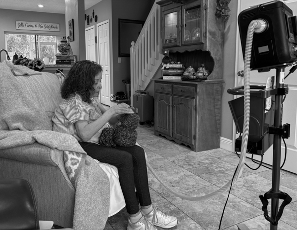 Woman attached to ventilator machine sitting in chair crocheting.