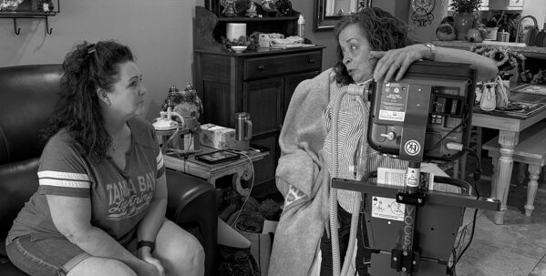 Maria Whitas sits in her living room on a ventilator for oxygen talking with another woman