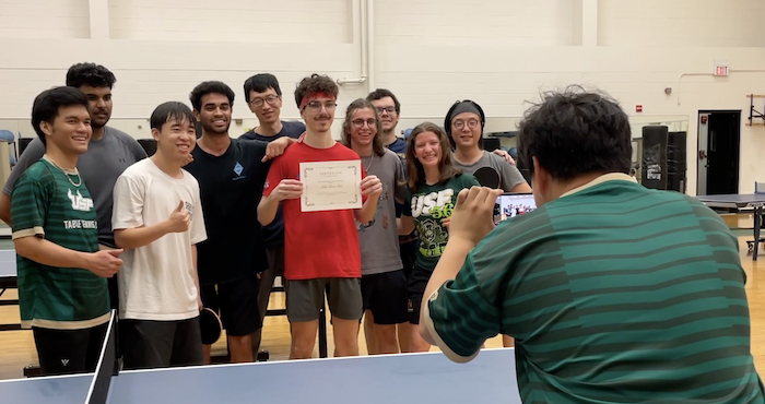 The USF Table Tennis poses together with a certificate in front of a man with a cell phone