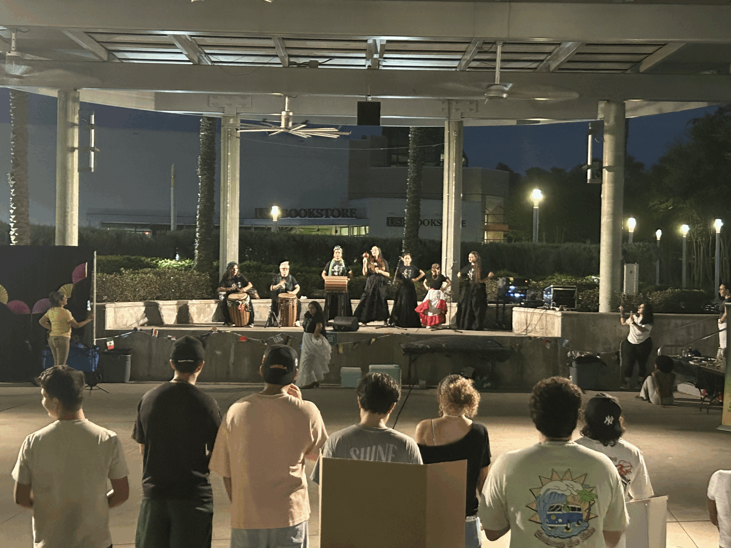 A group of performers on stage at night under a pavilion at the University of South Florida, dressed in traditional Latin American and Caribbean attire, while students and attendees watch from the plaza. Photo by Julia Lobo