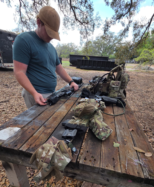 Outside environment, with trees and a dumpster in the background. A man is standing at a wooden table with various types of training and hunting gear. 