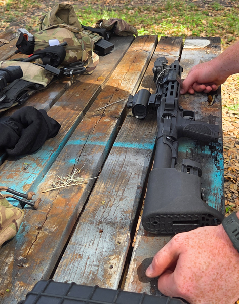 Outside environment. There is a wooden table with training gear and a gun on it. You can see a man's hands to the right side of the photo. 