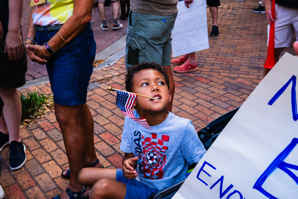 A photo of a kid with an American flag being held by his teeth amongst a crowd.
