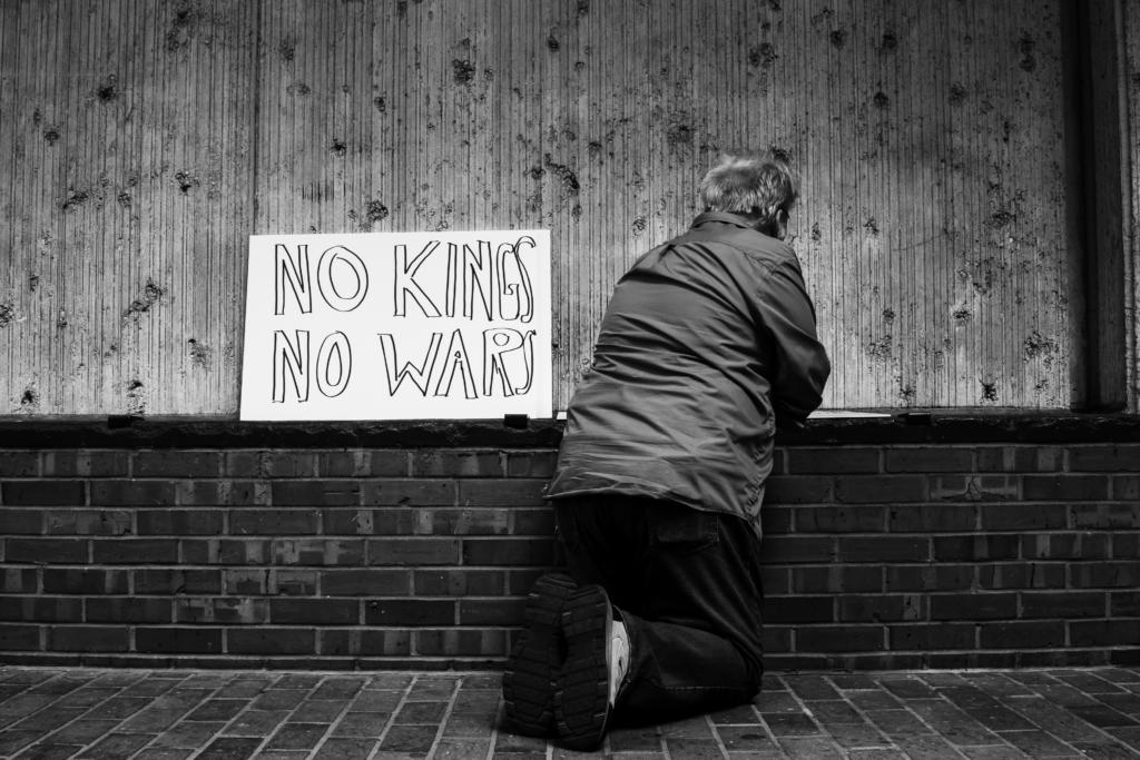 Black and white photo of man writing on a sign next to a sign that says "NO KINGS NO WARS"