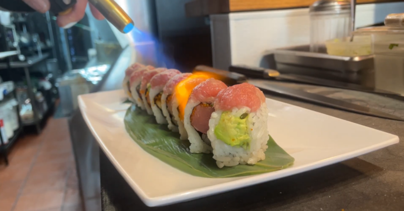 A chef in a restaurant kitchen uses a small torch to crisp a sushi roll