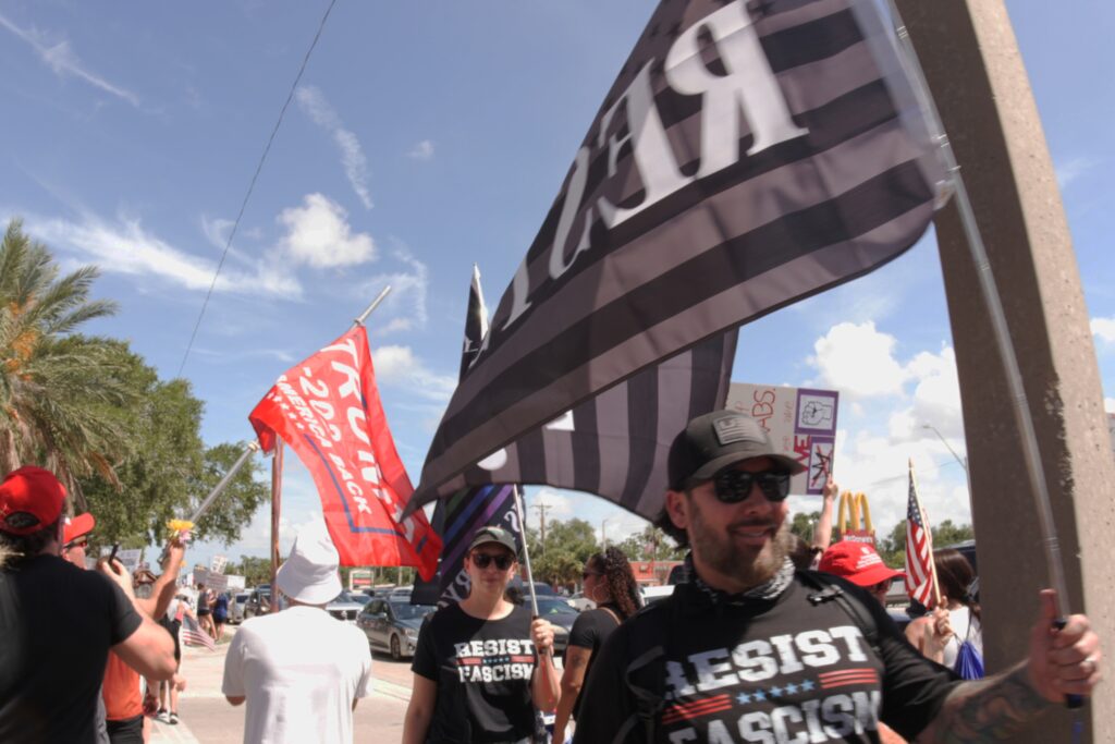 A photo of a person holding a flag that says "RESIST" crossing paths with a person holding a flag that says "TRUMP 2024".