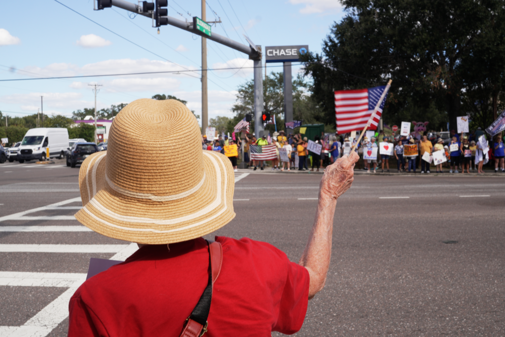 A photo of an older lady holding an American flag, across the street from a crowd of protestors holding signs.