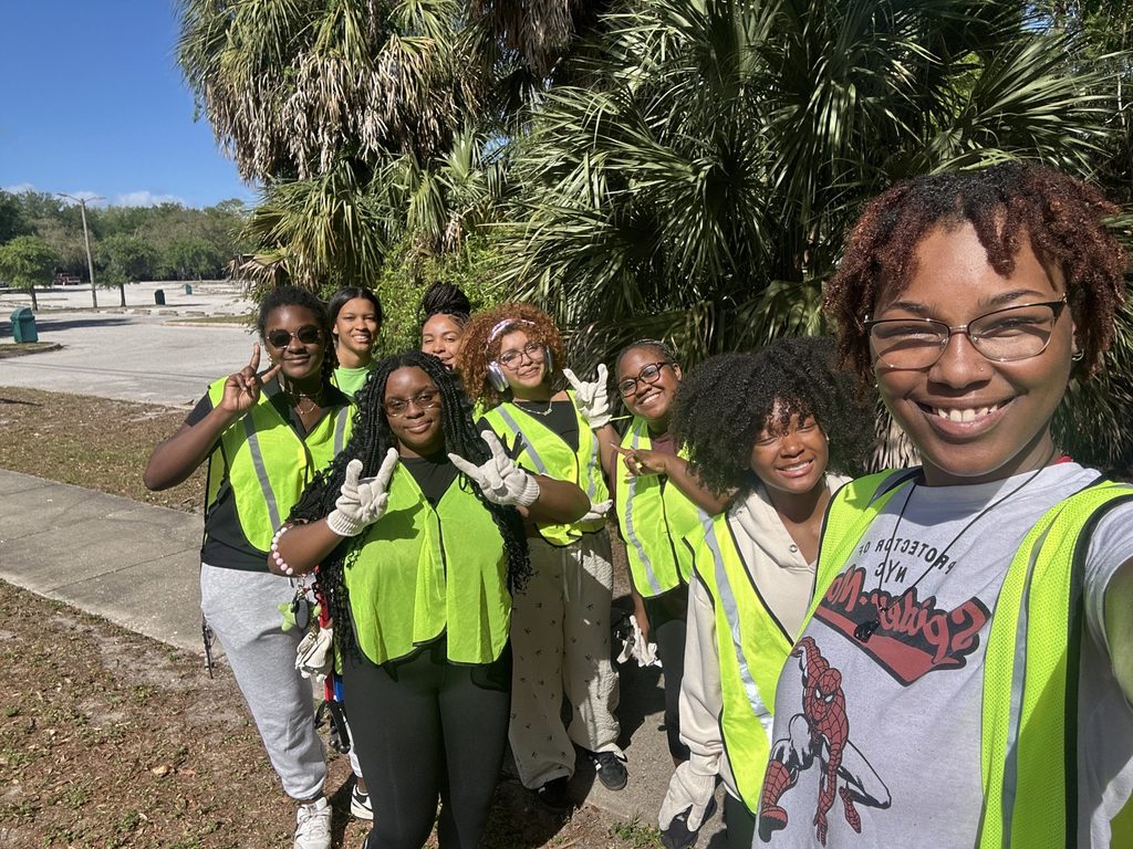 A selfie of seven young women smiling outdoors during a volunteer event. They wear neon-green safety vests and work gloves while posing closely in front of palm trees.