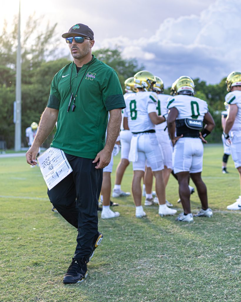 Chris Maragos wearing green Celtics gear walking across the football field in front of players in white and gold jerseys.