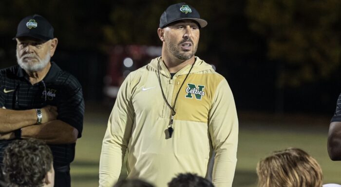 Football coach Chris Maragos talking to his team that is kneeling during a night football game.