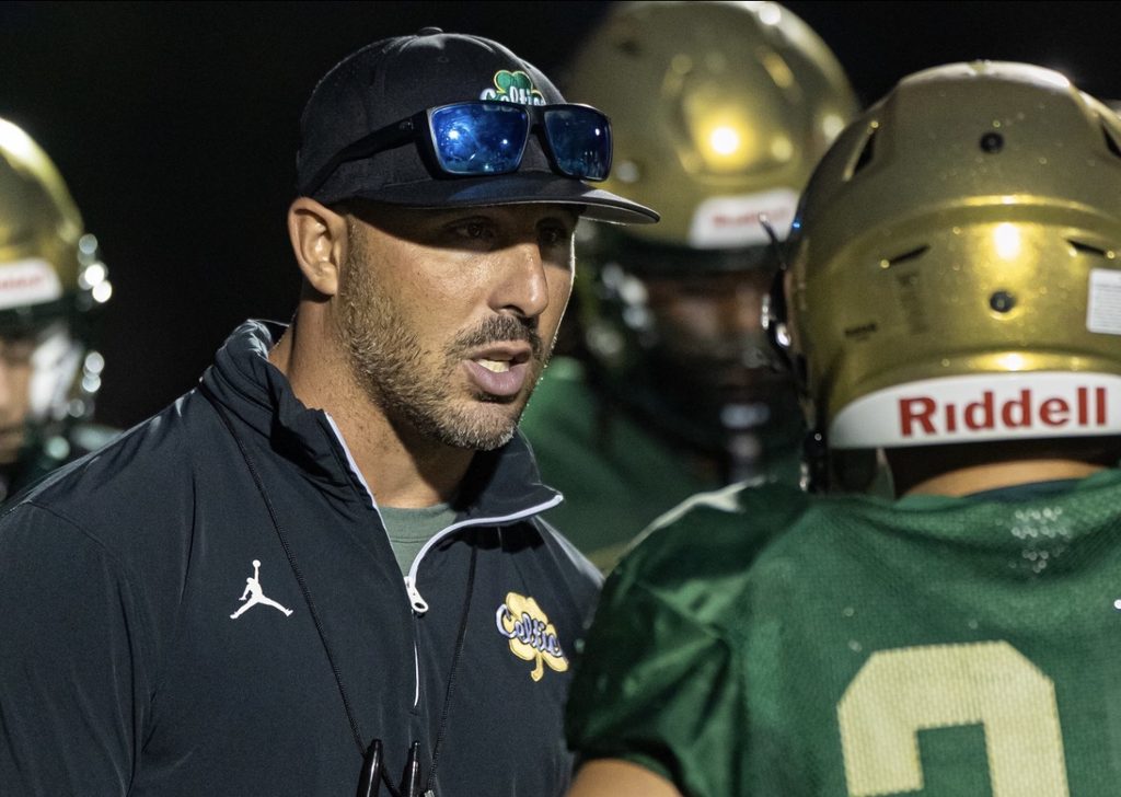 Football Coach Chris Maragos speaking to his player dress in a green jersey and gold helmet during a night game.