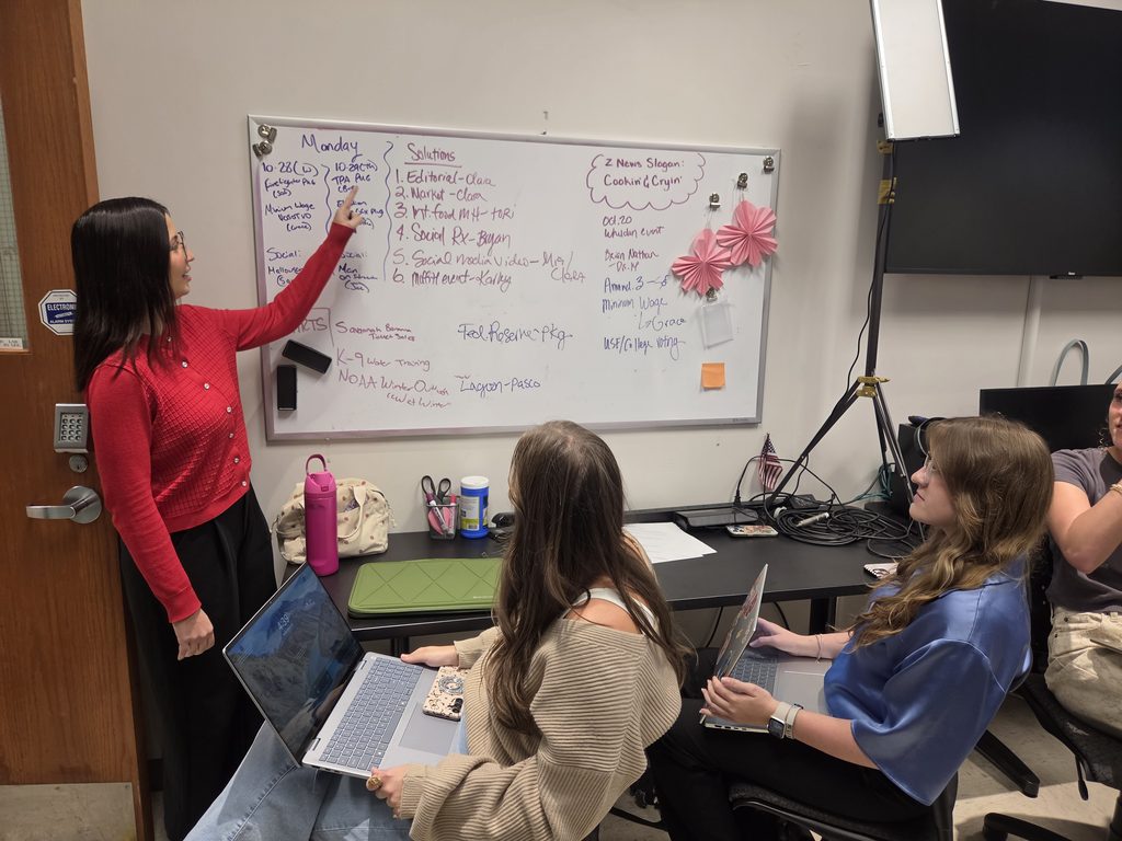 This is a photo with a white board with writing on it and a student in red pointing and speaking about what it listed on the board