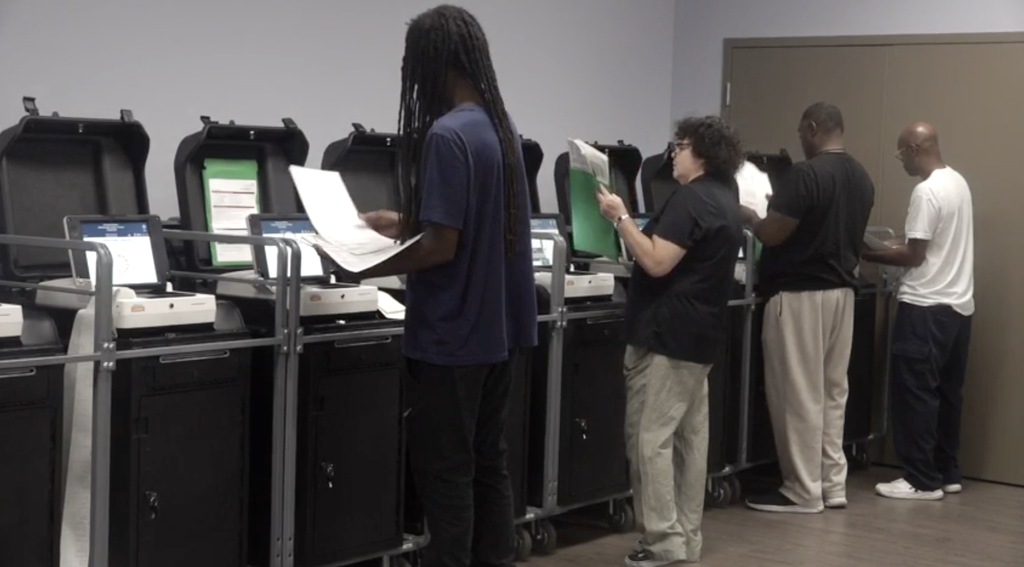 There is a man in the middle of a line of black voting machines, wearing a blue shirt looking at the voting sheet with a woman and a man to his left looking at a voting sheet as well. Another man in a white T-shirt is to the farthest left filing his voting sheet.