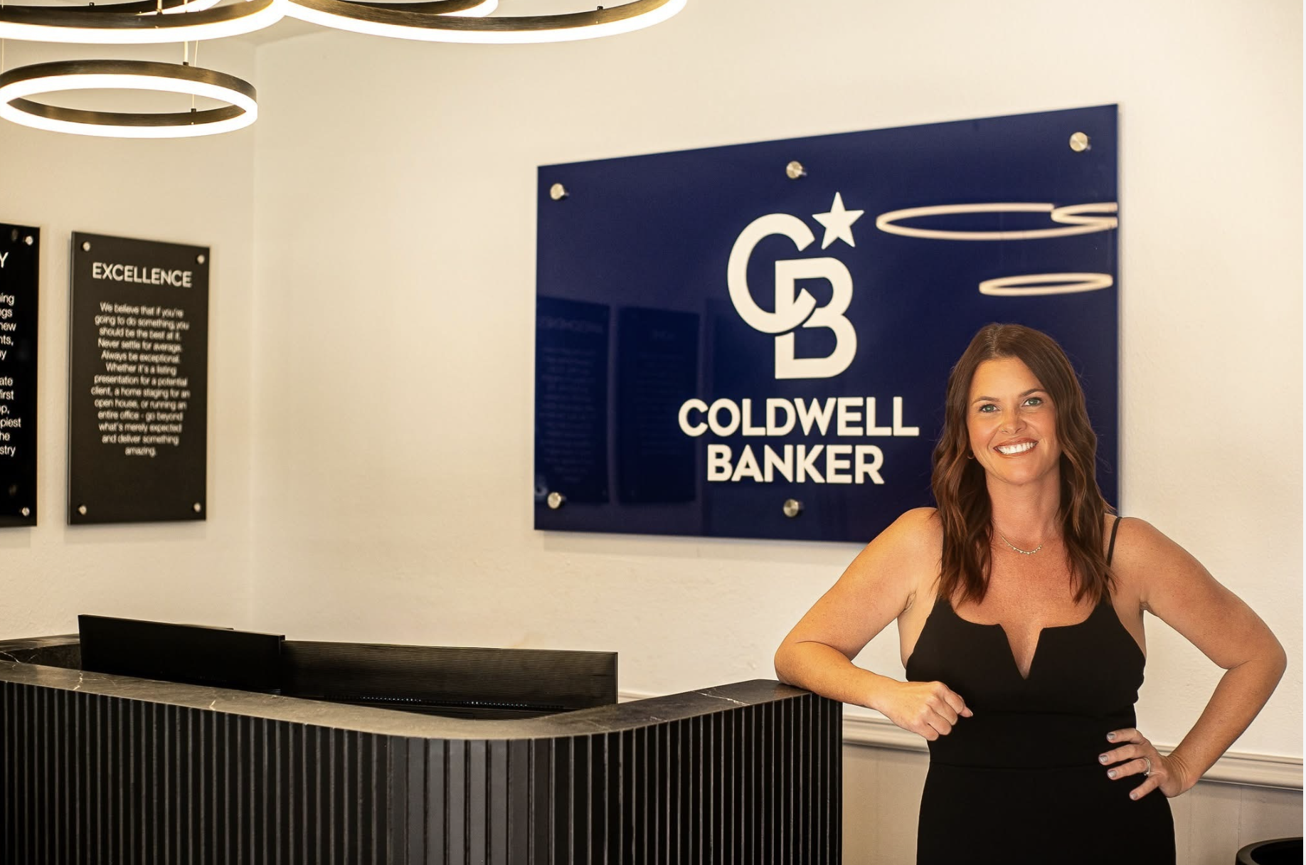 Realtor Chelsea Rose stands beside the front desk at a Coldwell Banker office in front of the company’s logo.