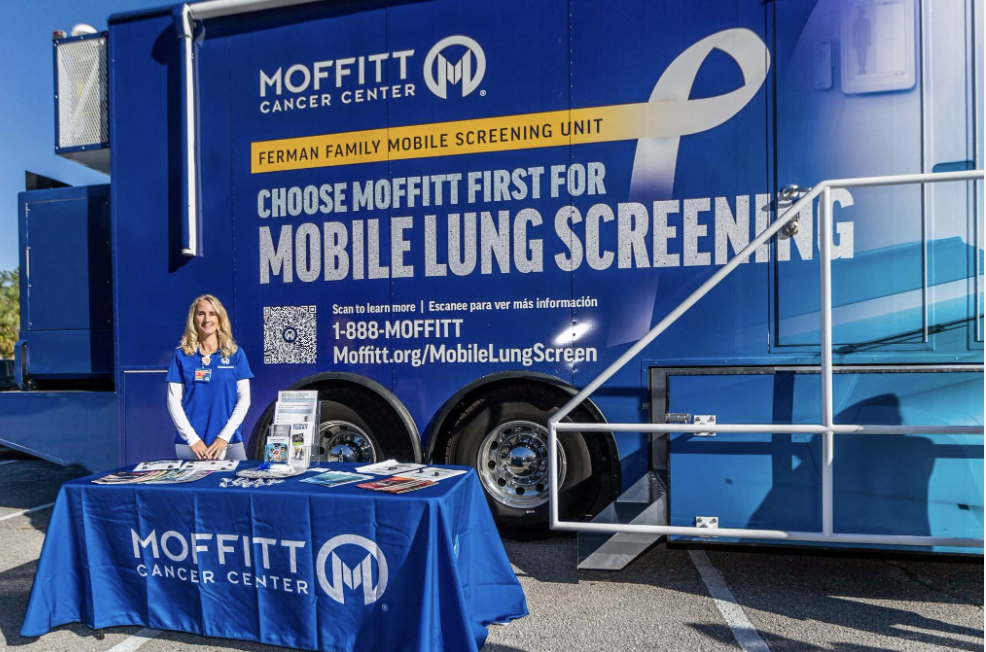 A Moffitt Cancer Center representative stands behind an information table in front of a blue mobile lung screening unit.