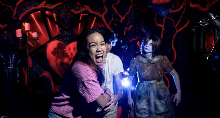 This shows a woman screaming with a character doll behind her in a haunted house setting